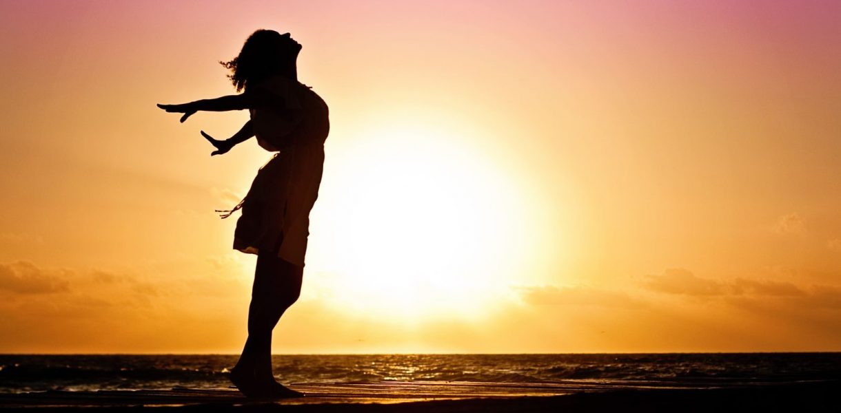 retour à soi se reconnecter Silhouette of a woman at the beach with arms outstretched against a vibrant sunset backdrop.