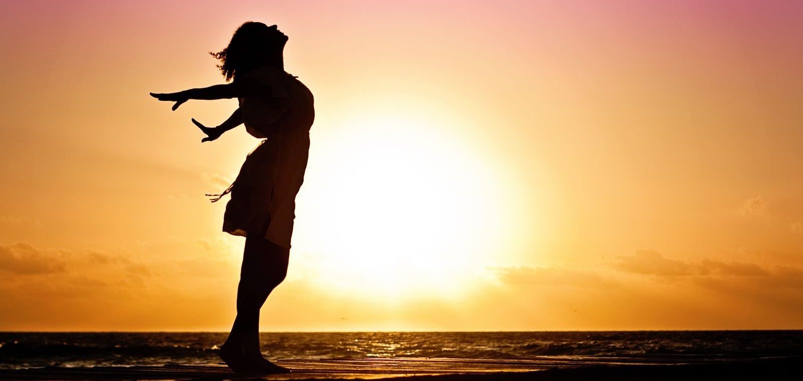 retour à soi se reconnecter Silhouette of a woman at the beach with arms outstretched against a vibrant sunset backdrop.
