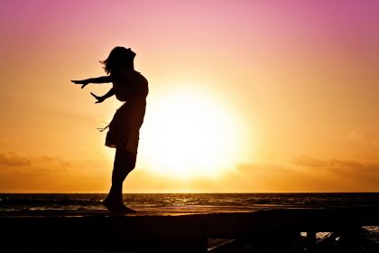 retour à soi se reconnecter Silhouette of a woman at the beach with arms outstretched against a vibrant sunset backdrop.