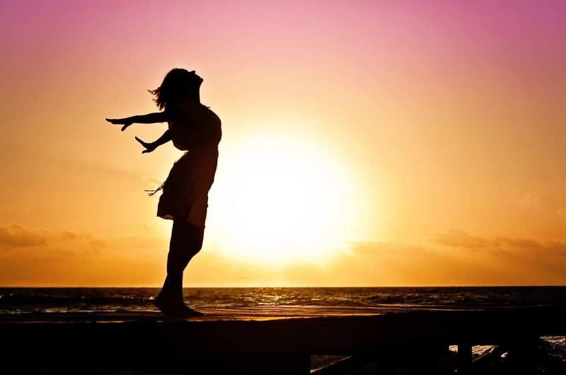 retour à soi se reconnecter Silhouette of a woman at the beach with arms outstretched against a vibrant sunset backdrop.